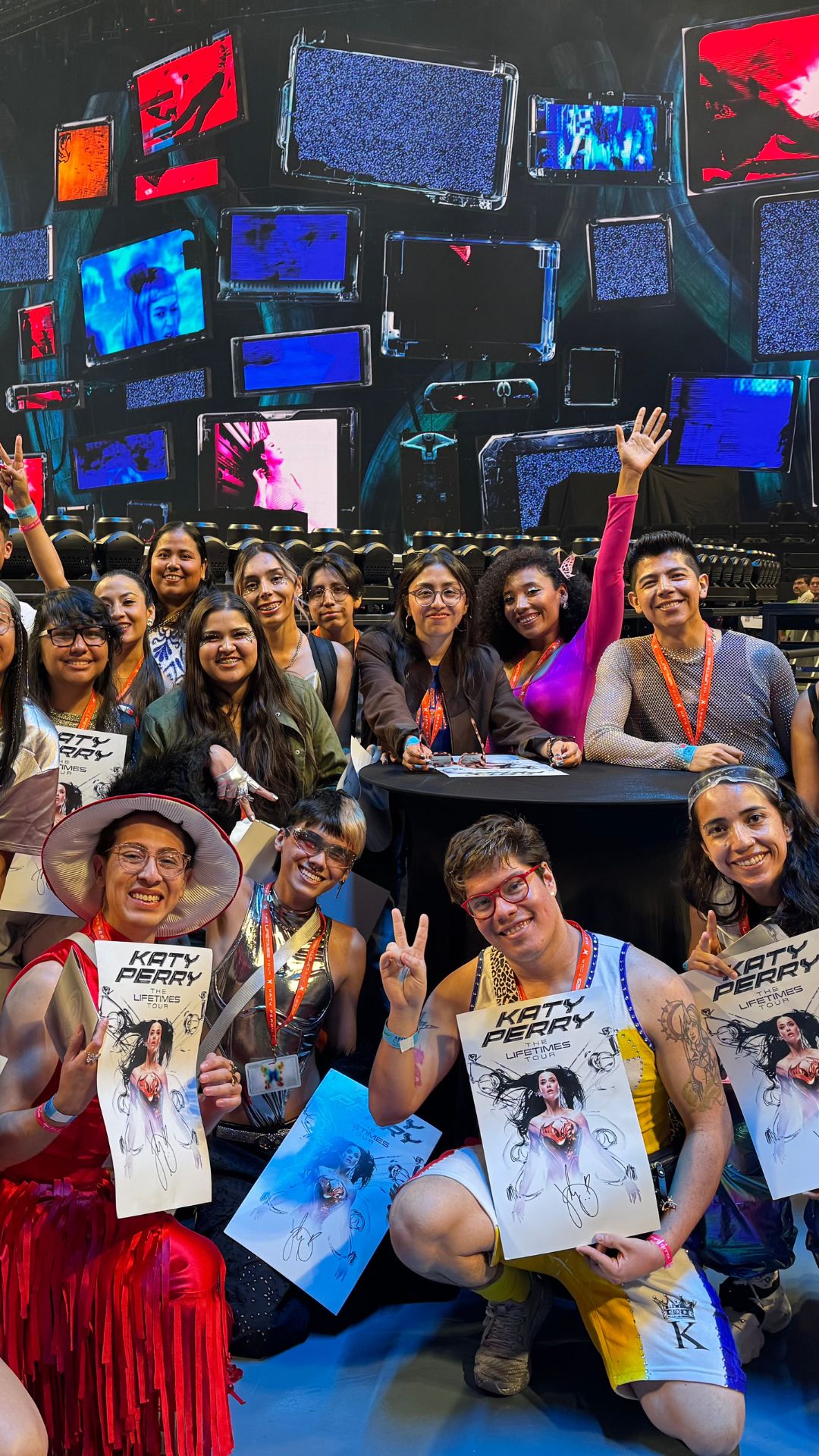 A group of VIP fans at Katy Perry’s 'The Lifetimes Tour' smile and pose on the arena floor, holding tour posters in front of a wall of colorful TV-screen visuals.